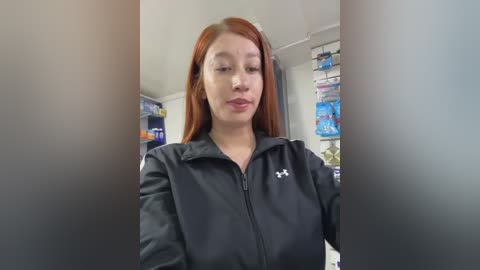 Video of a young woman with long red hair, wearing a black Under Armour jacket, standing in a pharmacy. Background features shelves of medicine and cosmetics.