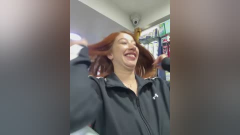 Video of a joyful woman with long red hair, wearing a black jacket, dancing in a brightly lit store with shelves of products in the background.