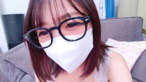 Video of an East Asian woman with shoulder-length brown hair, wearing black-rimmed glasses, a white face mask, and a light gray tank top, sitting on a gray couch with patterned pillows.