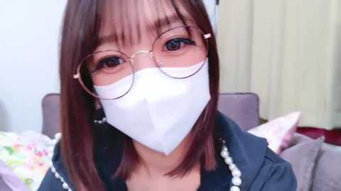 Media: Video of an Asian woman with shoulder-length brown hair, wearing round glasses, white surgical mask, and a dark jacket, standing indoors near a bed with floral pillows.