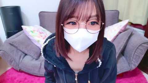 Media: Video of an Asian woman with straight, shoulder-length brown hair, wearing glasses, a white mask, and a black hoodie. She sits on a gray couch with floral pillows, in a room with a pink rug.