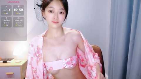 Media: Video of an Asian woman with fair skin and long black hair, wearing a pink floral kimono, revealing a pink bra. Background includes a clock, lamp, and gray curtains in a minimalist room.