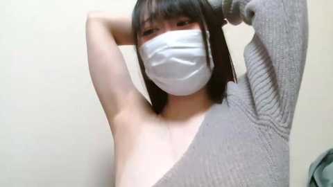 Media: A video of an East Asian woman with straight black hair, wearing a white medical mask and a grey ribbed sweater, raising her arms in a casual pose against a plain beige wall.