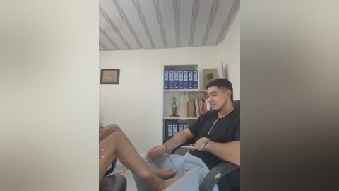 Media: Video of a young man in a black t-shirt and shorts, sitting in an office chair with crossed legs, looking at a woman's feet, in a sparsely furnished room with white walls, shelves full of binders, and a framed certificate.