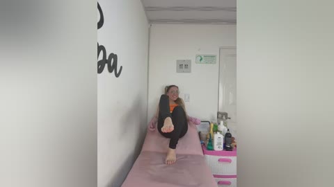 Media: A video of a young girl with long blonde hair in a white room, lying on a pink bed, wearing an orange shirt and black pants, surrounded by toys and a sink.