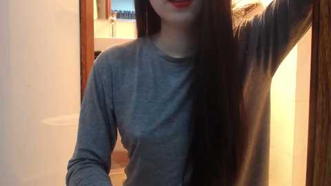 Media: Video of a young woman with long, straight black hair, wearing a light blue long-sleeve shirt, standing in a bathroom with a white sink and mirror in the background.