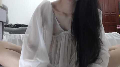 Media: Video of an Asian woman with long black hair, wearing a sheer, white blouse, sitting on a bed, legs crossed, in a dimly lit bedroom with a closed red door in the background.