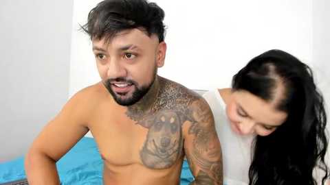 Media: Video of a shirtless South Asian man with a tattoo sleeve and beard, looking surprised, and a smiling Asian woman with long black hair, both on a blue bed.