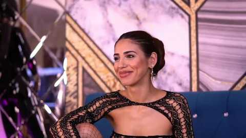 Media: Video of a confident woman with dark hair styled in a bun, wearing a black, intricate lace top with cut-outs, standing against a backdrop of metallic, geometric shapes in shades of blue and gold.