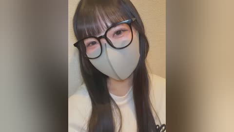 Video of an East Asian woman with long, straight black hair and bangs, wearing black-framed glasses and a beige surgical mask, standing against a beige wall, smiling slightly.