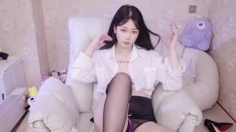 Media: A video of an Asian woman with long black hair, wearing a white blouse, black shorts, and thigh-high stockings, sitting in a plush chair in a pastel-colored room.