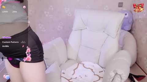 A video of a person wearing black shorts with pink floral print and a white, tufted leather chair. The room has a light pink floral wallpaper and a blue cushion.