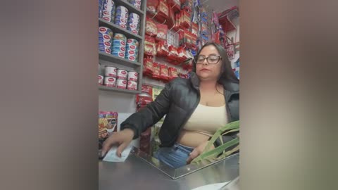 Media: Video of a middle-aged woman with glasses, dark hair, and medium skin tone, wearing a beige top and black leather jacket, reaching into a vending machine in a brightly lit convenience store.