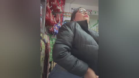 Video of a middle-aged, slightly overweight woman with glasses, wearing a puffy black jacket, standing in a grocery aisle. Red and green packaged snacks line the shelves behind her.