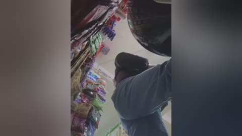 Video of a person in a surgical mask, leaning over colorful, neatly arranged snacks and drinks in a cluttered kitchen.