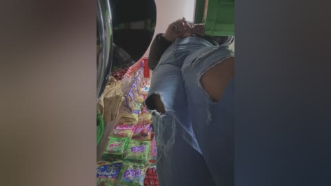 Media: Video of a person lying on a floor covered with brightly colored snack packages, wearing ripped jeans and a blue shirt, with a green wall and a dark background.