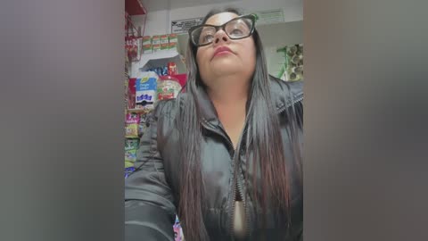 Media: Video of a plus-size woman with long black hair, wearing glasses, a black leather jacket, and a dog tag necklace, standing in a brightly lit convenience store aisle filled with colorful snacks.