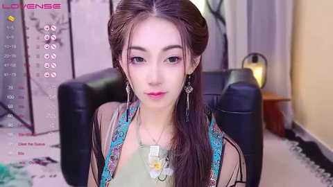 A video of a young woman with fair skin, brown hair, and large brown eyes, wearing a green top and colorful earrings, sitting on a black chair in a room with a gray floor and beige walls.