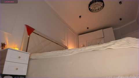 Media: Video of a modern bedroom with a white bed, beige walls, a red and white headboard, a white nightstand, a chandelier, and recessed lights.