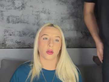 Video of a blonde woman with fair skin, wearing a blue shirt, looking shocked with her mouth open, against a gray textured wall.