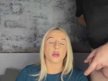 Video of a blonde woman with closed eyes and parted lips, wearing a blue shirt, sitting in a room with a textured, gray wall. A man's hand is visible in the foreground.