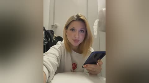 Media: Video of a blonde woman in a white sweater, holding a blue tablet, taking a selfie in a bathroom.