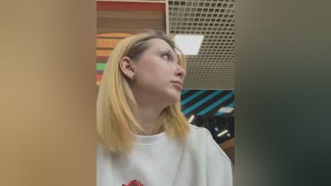 Media: Video of a young woman with shoulder-length blonde hair, wearing a white T-shirt, standing in a modern, brightly lit room with geometric patterns on the walls and a grid ceiling.