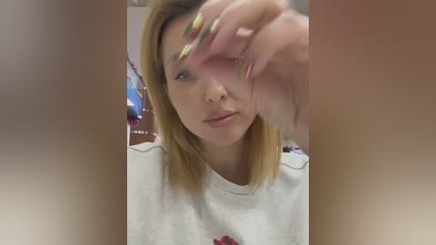 Media: A video of a young Asian woman with shoulder-length blonde hair, light skin, and wearing a white T-shirt, being touched on her face by another hand with long, multicolored nails. Background is blurred, indoor setting.