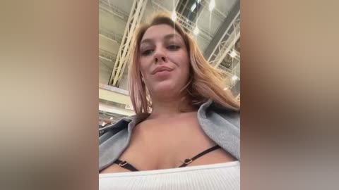 Media: Video of a young woman with light skin, long blonde hair, wearing a white tank top and grey hoodie, standing in a modern, industrial building with high ceilings and metal beams.