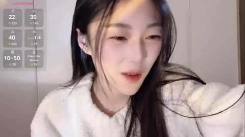 Media: Video of a young East Asian woman with long black hair, fair skin, and light makeup, wearing a white fluffy robe, indoors.