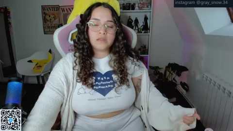 A video of a plus-sized woman with curly hair, wearing a yellow hat and a heart-patterned crop top, sitting in a gaming chair.