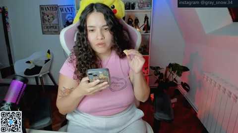 Video of a plus-size woman with curly hair, wearing a pink t-shirt and grey pants, eating a cookie, sitting on a pink gaming chair. Background includes a wall with posters, plants, and a desk with a phone charger.