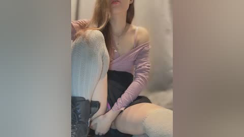 Video of a slender, fair-skinned woman with long brown hair, wearing a lavender off-shoulder top and black skirt, sitting on a couch, legs crossed, in a dimly lit room.