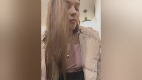 Media: Video of a young woman with long, straight, light brown hair, wearing a pink top, beige puffer jacket, and a heart pendant necklace, sitting in a dimly lit room.