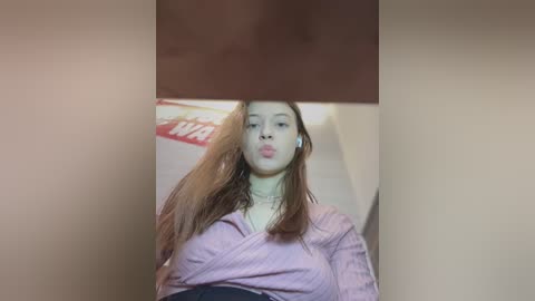 Video of a young woman with long, wavy brown hair, wearing a light pink striped shirt, making a pouty face. Background includes a blurred red and white \"Wawa\" sign.