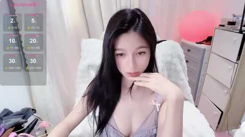 Media: Video of an East Asian woman with long black hair, fair skin, and a slender figure, wearing a lacy white bra, sitting on a bed with white bedding in a softly lit room.