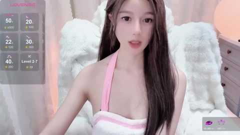 Media: Video of a young East Asian woman with long, straight brown hair, fair skin, and a slender physique. She wears a pink halter top, sitting on a white fluffy bed, surrounded by soft lighting.