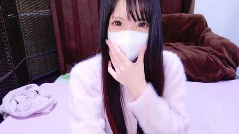 A video of a young East Asian woman with long black hair, wearing a white mask, a white fluffy jacket, and lying on a bed, surrounded by dark brown curtains and a white blanket.