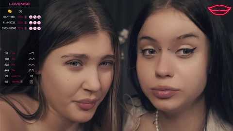 Video of two young women with light skin, dark hair, and makeup, possibly in a live video feed, set against a dark background.