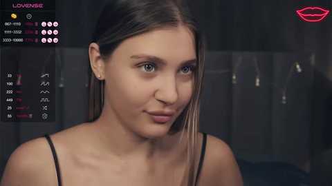 A video of a young woman with fair skin and straight, light brown hair, wearing a black spaghetti strap top. The background features a dimly lit room with a mirror and a pink lipstick mark.