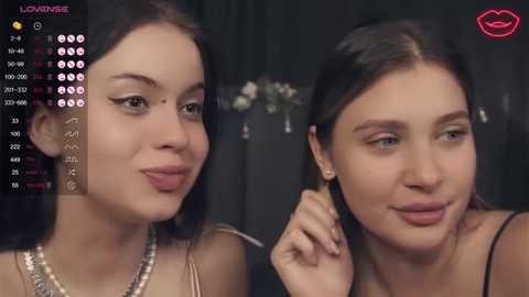 Video of two young women with light skin, dark hair, and makeup, smiling and holding hands; background shows a black curtain with a pink lipstick logo.