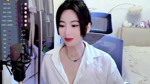 Media: Video of a young East Asian woman with short black hair, wearing a white button-up shirt, sitting at a desk with a microphone, in a cluttered bedroom with a blue chair and stuffed toys.