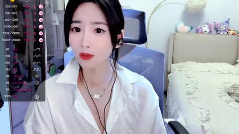 Media: Video of a young East Asian woman with fair skin, black hair in a bun, wearing a white shirt and red lipstick, seated in a room with a bed, stuffed toys, and a computer screen.