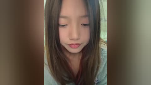 Media: A video of a young Asian girl with long, straight brown hair, fair skin, and light pink lipstick, gazing downward with a contemplative expression. She wears a light gray sweater.