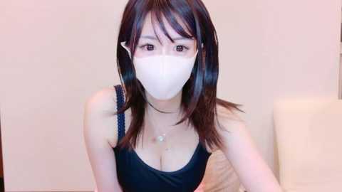 Media: Video of an East Asian woman with long black hair, wearing a white face mask, black sleeveless top, and a delicate necklace, sitting on a beige couch.