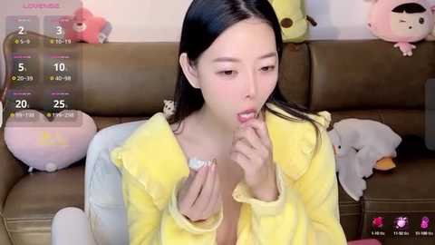 Media: Video of an East Asian woman with long black hair, wearing a yellow robe, eating a white snack, surrounded by plush toys on a brown couch.