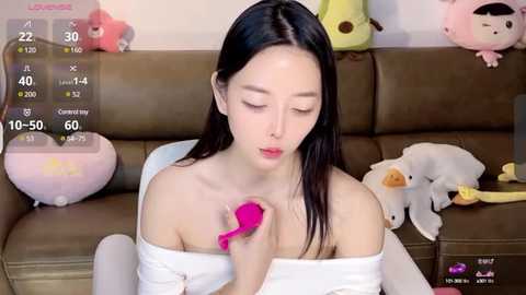 Media: Video of an East Asian woman with long black hair, wearing an off-shoulder white top, using a pink vibrator. Background includes a brown couch with plush toys, and a smartphone screen displaying \"Love Live!\" in the top left.