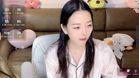 Media: A video of a young East Asian woman with long black hair, fair skin, and a slender physique, wearing a white robe, sitting on a brown leather couch. Soft toys are scattered around her.