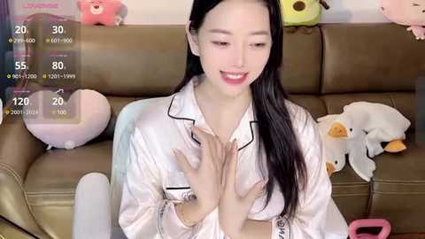 Media: Video of an East Asian woman with long black hair, fair skin, and small breasts, wearing a white satin robe, sitting on a brown leather couch. Background includes plush toys, a digital clock displaying 30\u00b0C, and a pink cup.