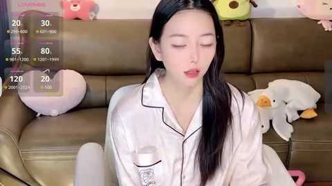 Media: Video of a young Asian woman with long black hair, wearing a white pajama shirt, sitting on a brown leather couch surrounded by stuffed animals.